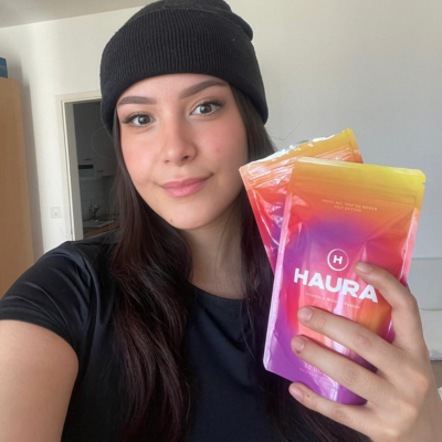 Woman in beanie holding colorful HAURA skincare products.