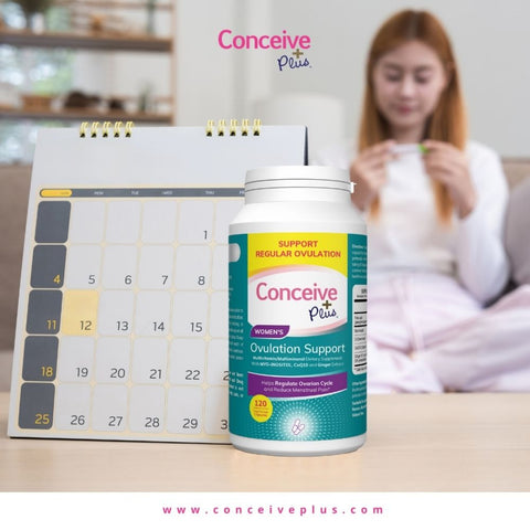 A woman in the blur background looking at a pregnancy strip while sitting at a sofa and a Conceive Plus Ovulation support bottle placed in front of a calendar
