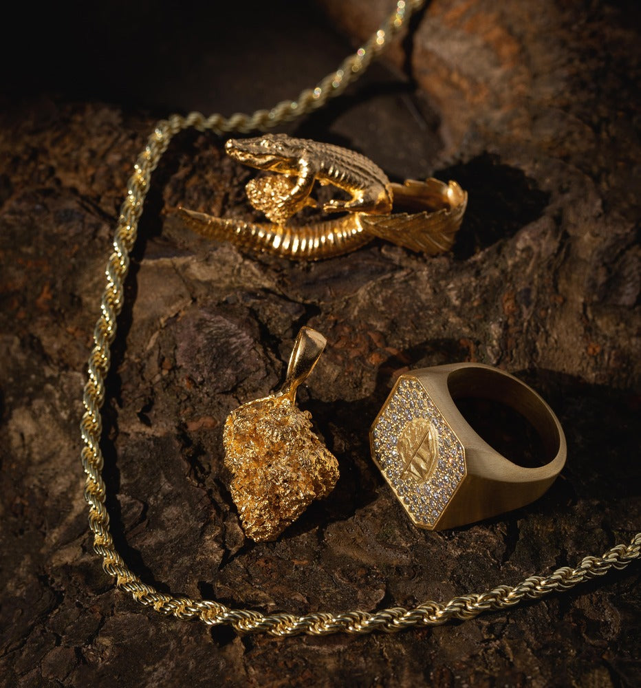 Gold jewelry: a chain, nugget pendant, crocodile brooch, and ring displayed on textured surface.