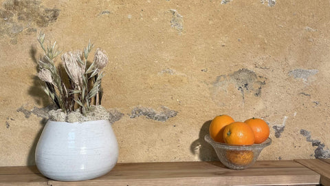 Composition florale avec un support en poterie posé sur une tablette en bois à côté de quelques oranges dans une panière de fruit.