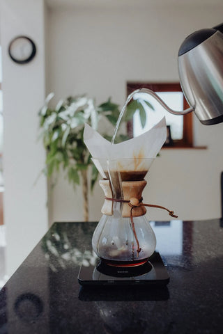 Chemex coffee maker on a scale on a black table being filled by a gray csine neck carafe.