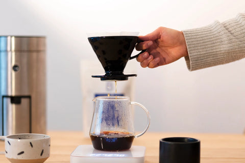 Wooden table with a v60 coffee maker on a weight and a hand lifting the dripping filter after coffee extraction.