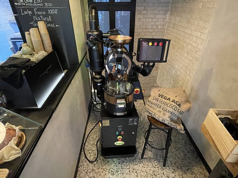 Coffee roaster inside a premises, on the left a coffee maker and on the right a stool with a sack of coffee.