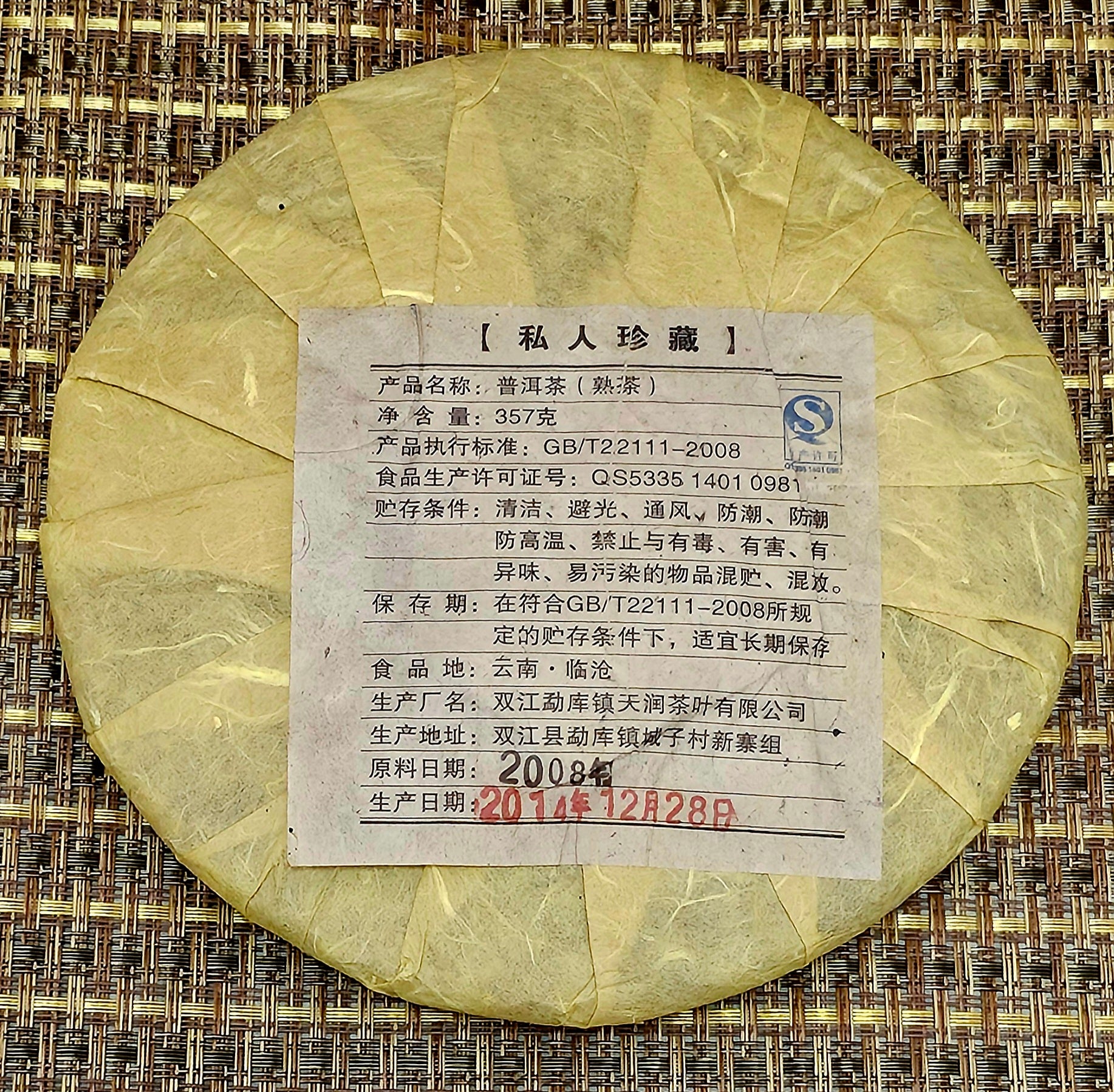 A round package wrapped in yellow paper with a Chinese label featuring product information rests on a woven mat, containing Mansong Prince Mountain Tribute Tea - Wujin No.1 烏金壹號 (Puerh Ripe Tea) from ZenTea Shop.