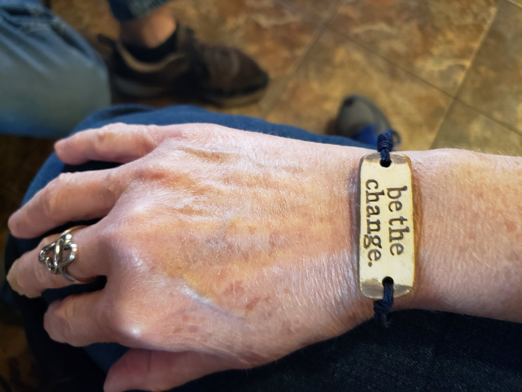 Image depicts a "be the change" bracelet on a woman's wrist. 