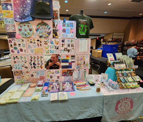 My artist alley table at Fursonacon 2023, with a wall of cube panels displaying stickers, prints, and buttons, a set of racks holding enamel pins and sticker books, and stationery and prints laying out on the table.