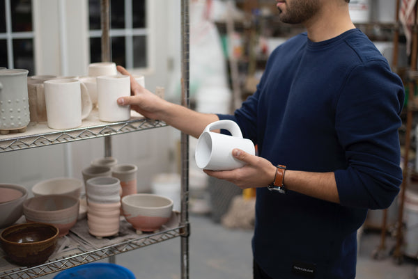 Man holding the ceramic CURVD mug prototype at a ceramic studio next to a shelf of mug prototypes.