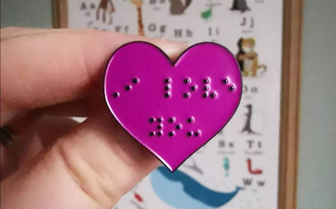 A fuchsia pink heart shaped pin with I love you in braille.