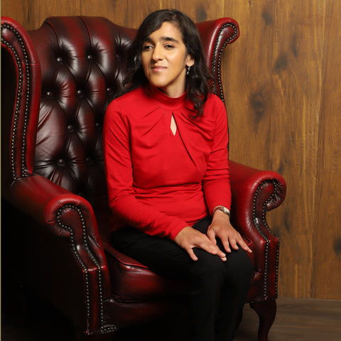 Zubbe, a woman of colour sitting in a chair, wearing a bright red top and black trousers.