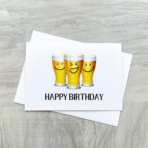 An A6 landscape white greetings card with three beer pint glasses with different cheeky faces, beneath that it says HAPPY BIRTHDAY in large print. There is a white envelope beneath the card.