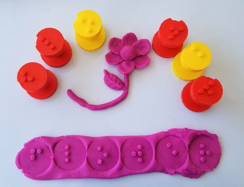 Six red and yellow braille play dough stampers with purple play dough with letters imprinted and flower made from the play dough.