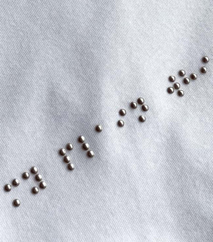 A close up a white/grey t-shirt with braille letters made with beads, the braille we can see reads my plain whi and then it cuts off.