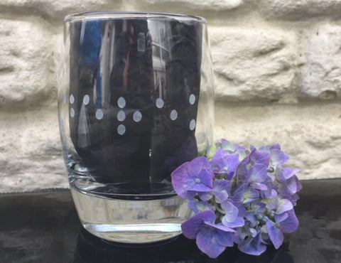 A whisky glass with braille is only part visible, the letters we can see are berek. There is a purple flower next to the glass.