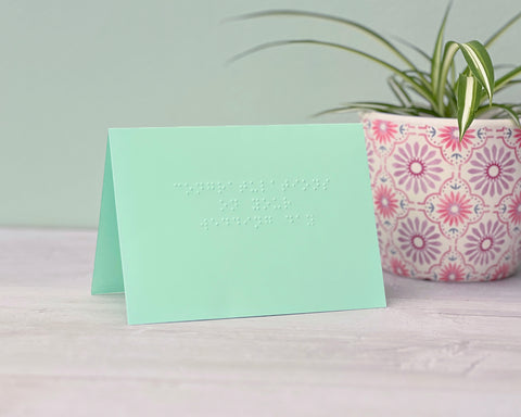 A pastel green landscape card saying congratulations on your wedding in lower case braille. There is a plant pot to the right.A