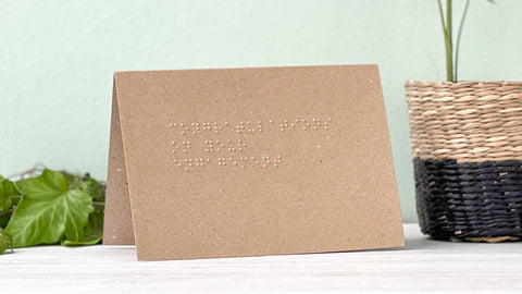 A kraft brown landscape card with congratulations on your engagement written in braille, there is foliage to the left and a plant on the righ