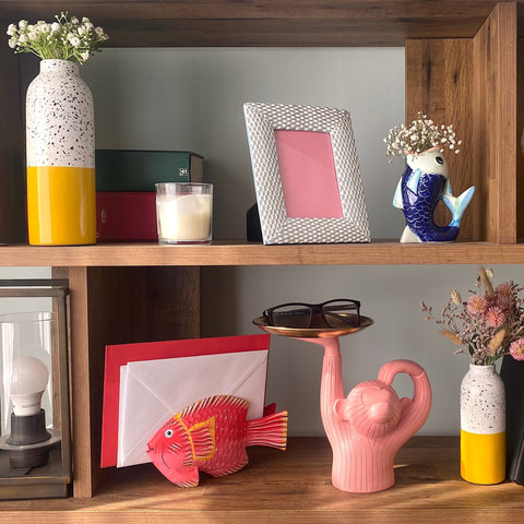 wooden shelving unit against a blue wall. the shelving unit has multiple small home decor items on such as a blue picture frame, a pink fish shaped letter rack and yellow tall vases.