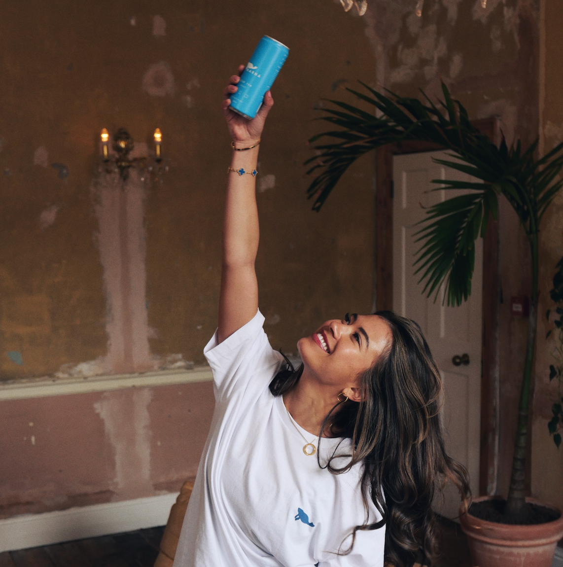 Woman smiling and holding up a blue can in a room with a plant.