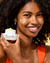 A model smiles while holding an open jar of belif Aqua Bomb Brightening Vitamin C Cream with some cream on her cheek