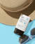 A white tube of belif The True Cream - Aqua Bomb Sunscreen lying on a light blue surface among dark sunglasses with light blue frame and a straw hat.
