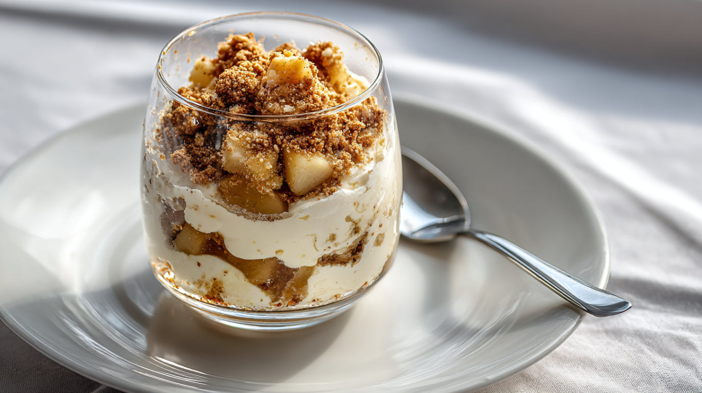 Bratapfel-Dessert im Glas mit Spekulatius-Crunch, Apfelstücken und Creme, serviert auf Teller mit Löffel.