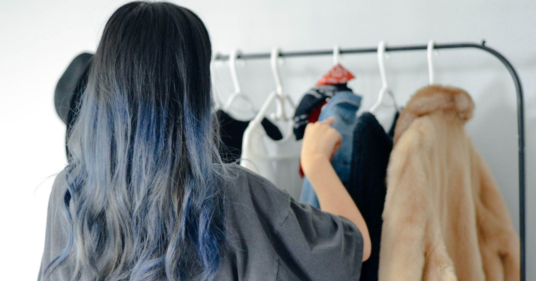 Mulher virada de costas, de cabelo azulado e uma camiseta cinza e larga, organiza uma arara de roupas