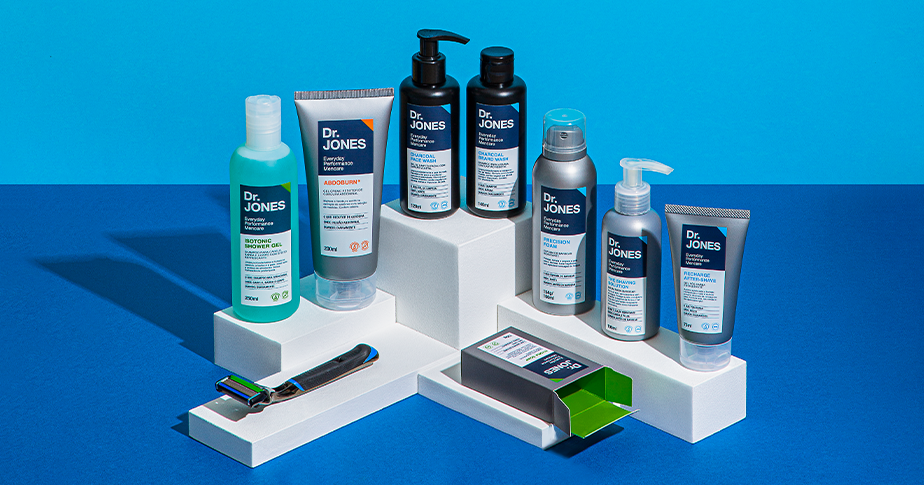 Um display branco ao centro da imagem mostra os produtos da Dr. JONES. O display está sobre uma mesa azul escuro e o fundo da imagem é azul claro.