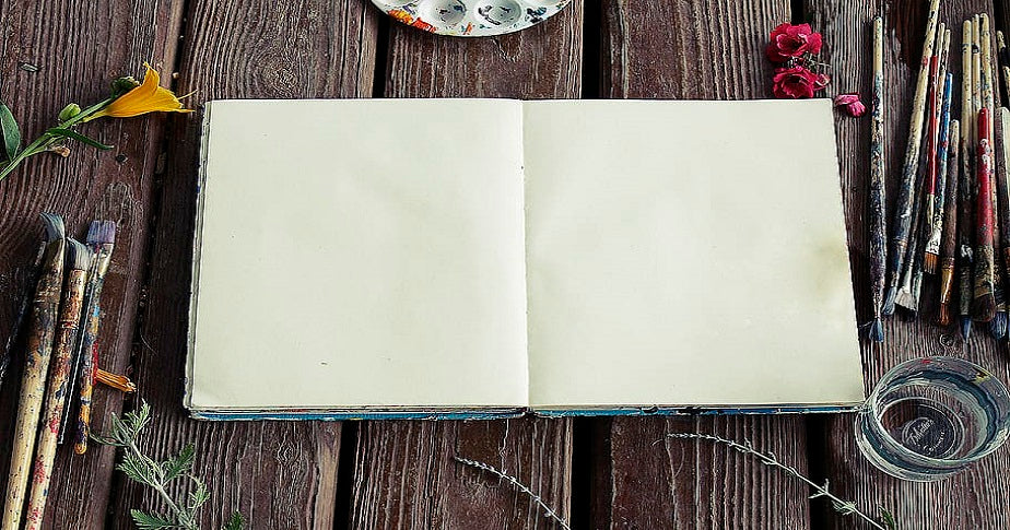 caderno aberto nas páginas em branco com pincéis sujos de tinta ao redor sobre mesa rústica de madeira e outros elementos