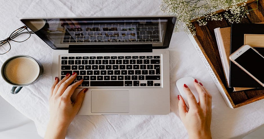 flatlay de uma mulher trabalhando em casa no laptop com xícara de chá e óculos do lado esquerdo e flores, celular, livros e cadernos do lado direito