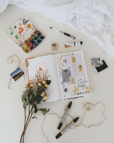flatlay de um caderno com colagens, estojo de tintas de aquarela usadas, fotos polaroid, flores e outros elementos artísticos