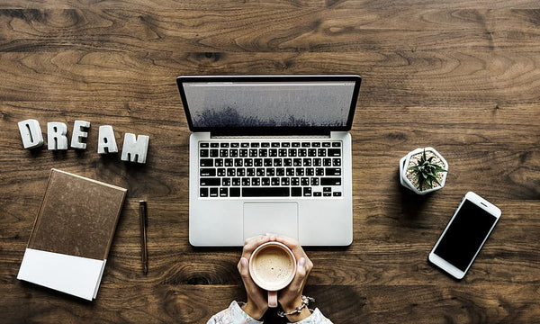flatlay de uma mesa trabalho com laptop  e uma xícara de café ao centro, a palavra dream, um caderno e uma caneta à esquerda e um vaso de planta e um smartphone à direita