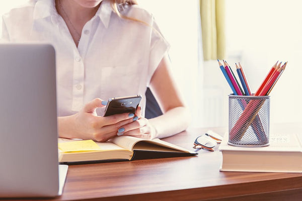 Como trabalhar pela internet: mulher teclando no smartphone em mesa trabalho com laptop, agenda, óculos, livro e porta-lápis