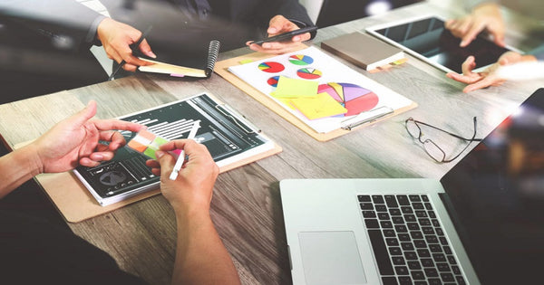 pessoas em uma reunião de trabalho sobre trade marketing ao redor de uma mesa com tablets, gráficos, agendas e laptop