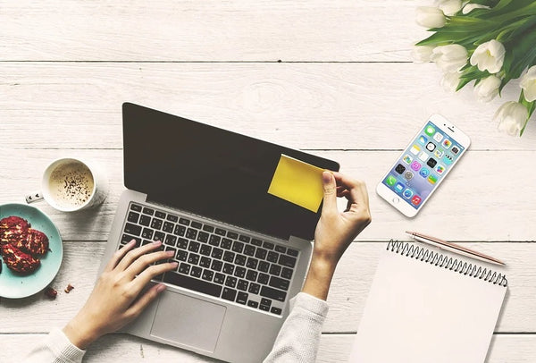 mulher trabalhando no laptop em mesa com doces e café de um lado com celular e caderno do outro