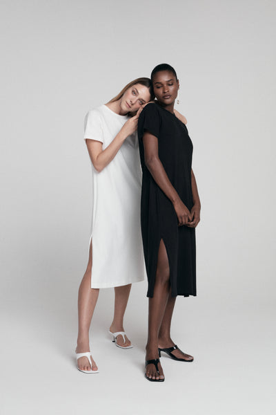 Foto sobre moda atemporal mostra duas pessoas de pé, uma usando um vestido branco e a outra com um vestido preto. A foto é de corpo inteiro e a pessoa à esquerda está com a cabeça e as mãos sobre os ombros da pessoa à direita.