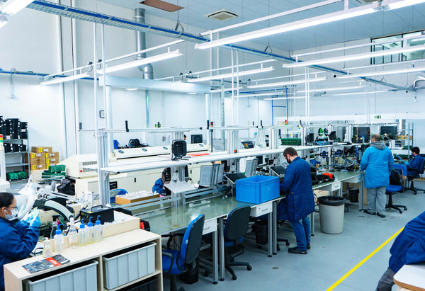 Foto mostra diversos funcionários diante de uma bancada de uma fábrica. Os funcionários estão usando um uniforme azul.