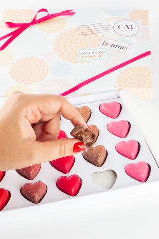 Foto de artigo sobre o que vender na Páscoa 2022 mostra uma caixa de bombons em formato de coração, com a embalagem da CAU Chocolates. Há uma mão de uma pessoa pegando um dos bombons da caixa.