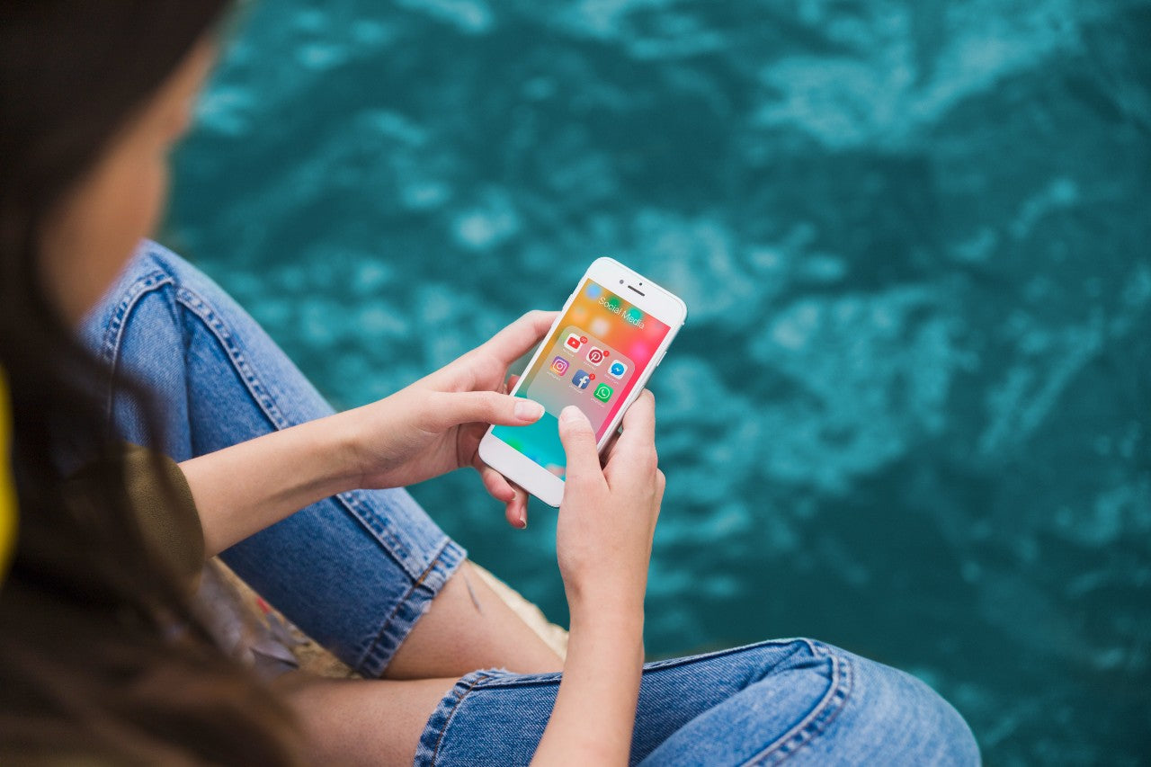 Uma mulher jovem, sentada de pernas cruzadas no chão, segura um smartphone com as duas mãos. Na tela do smartphone, há ícones de redes sociais, como o Instagram.