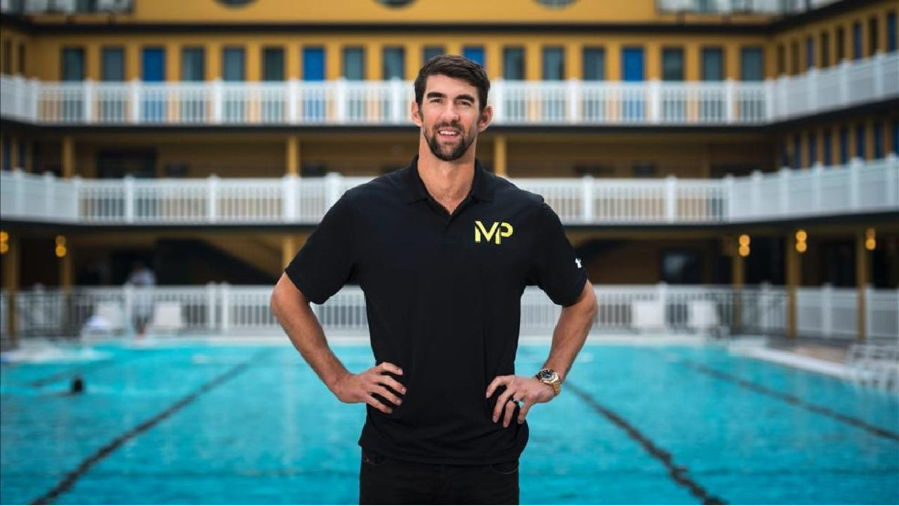 Michael Phelps com as mãos na cintura, sorridente, com uma piscina olímpica ao fundo.