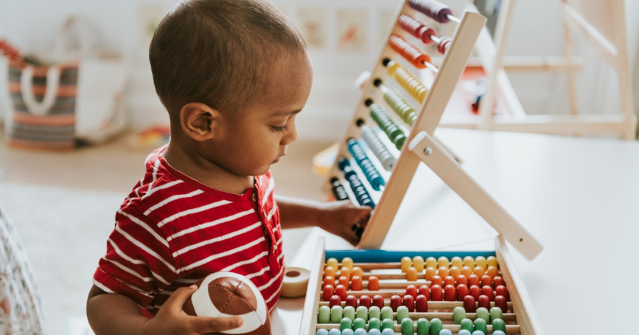 Brinquedos para crianças e bebês são a pedida certa para quem está buscando o que mais vende hoje. A fotografia mostra uma criança pequena, vestindo uma camisa listrada, observando e tocando em brinquedos de madeira.