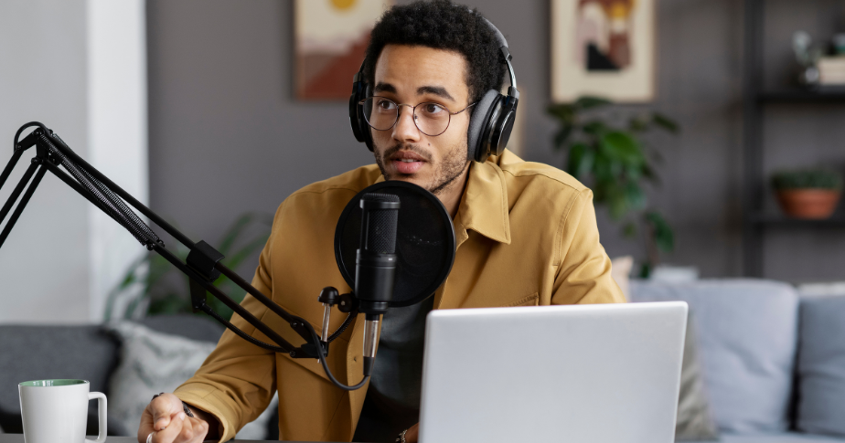 Microfones para podcast estão entre o que mais vende hoje. A fotografia mostra um homem, bastante jovem, vestido de forma mais formal e sentado em frente a um notebook. Ele está com fones profissionais e um microfone de podcast.