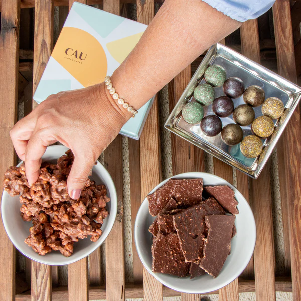 Foto publicitária da CAU Chocolates para artigo sobre ideias para Páscoa mostra duas tigelas sobre uma mesa de madeira, cada uma com pedaços de chocolate diferentes. Há também uma caixa de bombons aberta com o logo da CAU Chocolates, e há uma mão de uma pessoa pegando um pedaço de chocolate da tigela.