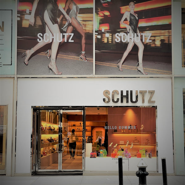 Fachada da loja da Schutz é branca, com vitrines e portas de vidro. O logo da Schutz está no canto superior direito da fachada, e acima há dois pôsteres com modelos usando os produtos da loja.