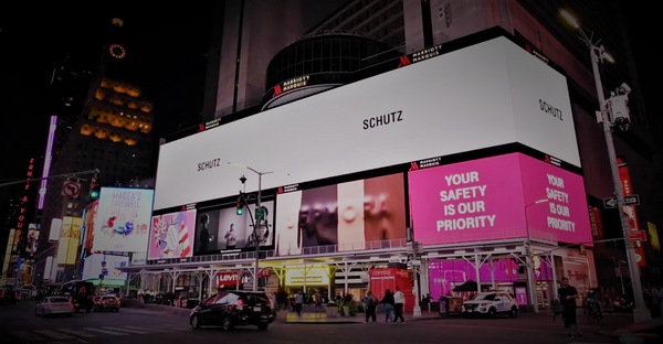 Foto tirada à noite de uma loja de esquina na Times Square, em Nova York. A fachada da loja é toda coberta por painéis luminosos, com anúncios de diversas marcas. No painel superior, em branco, há o nome da loja Schutz.