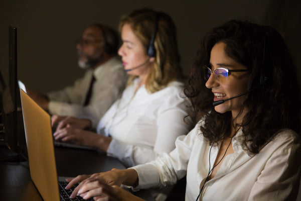 Foto mostra três pessoas sentadas lado a lado, diante de laptops e com headphones, trabalhando em atendimento ao cliente.