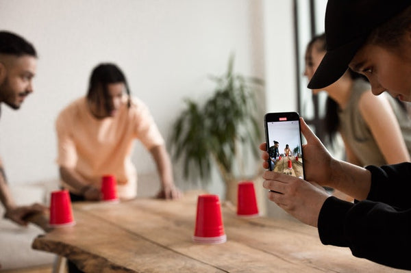Foto mostra quatro amigos ao redor de uma mesa retangular. Sobre a mesa, há quatro copos plásticos na cor vermelha. Três dos amigos estão jogando um jogo com os copos, enquanto a quarta pessoa segura um celular e filma a interação.