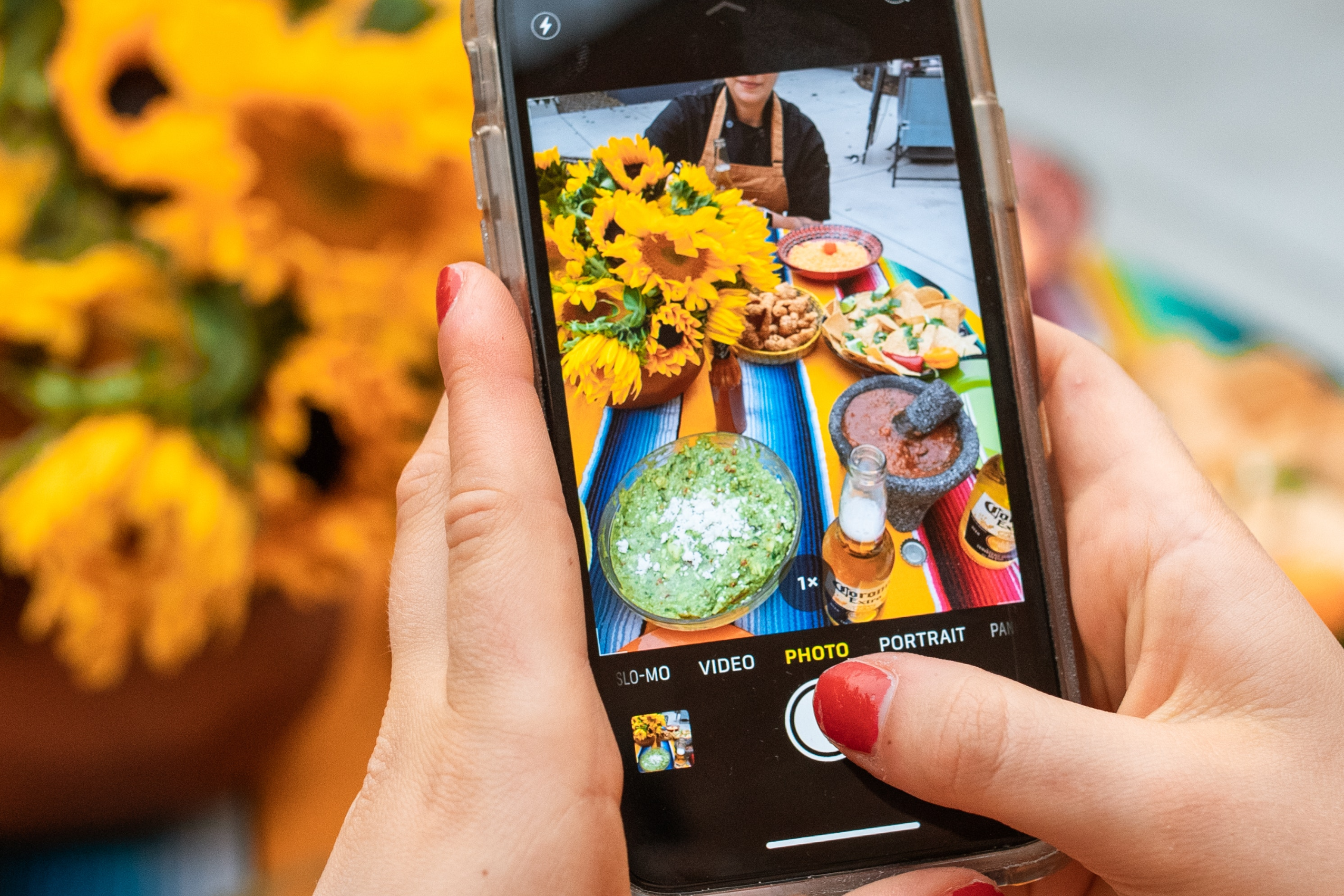 A imagem mostra, ao fundo, uma mesa arrumada com girassóis e comidas. No primeiro plano, celular fotografa a cena.