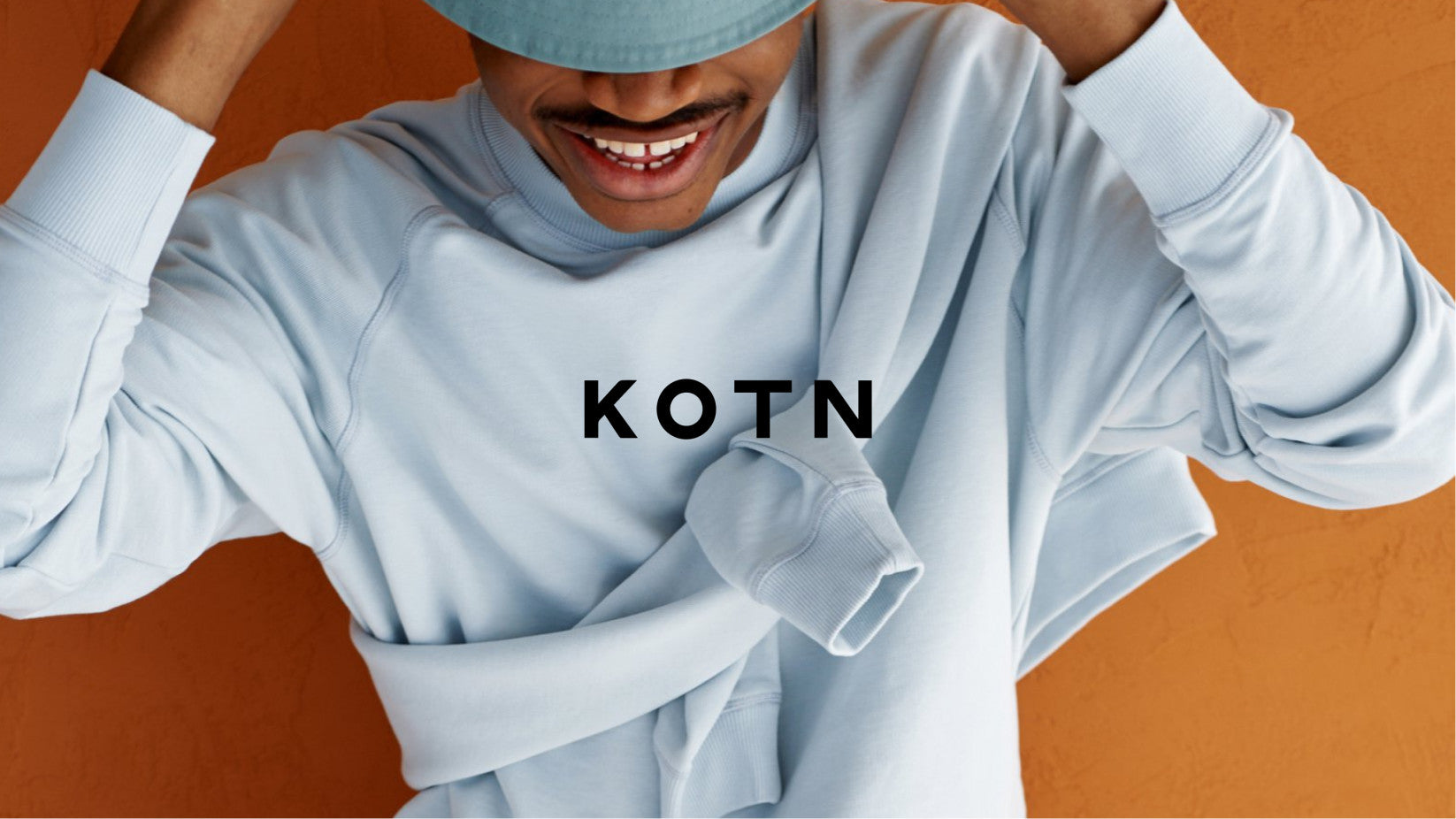 Homem com camiseta de manga comprida branca contra fundo laranja e o logo da Kotn em destaque na frente da imagem.