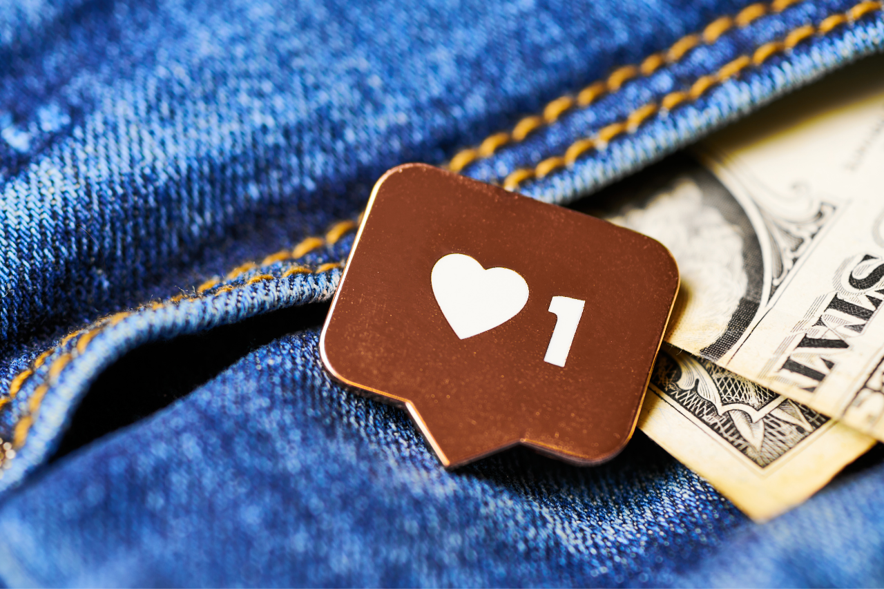 Sob um fundo de jeans, que parece o bolsode uma calça, estão duas notas de dólares e um pin de enamel com o símbolo de uma curtida do Instagram