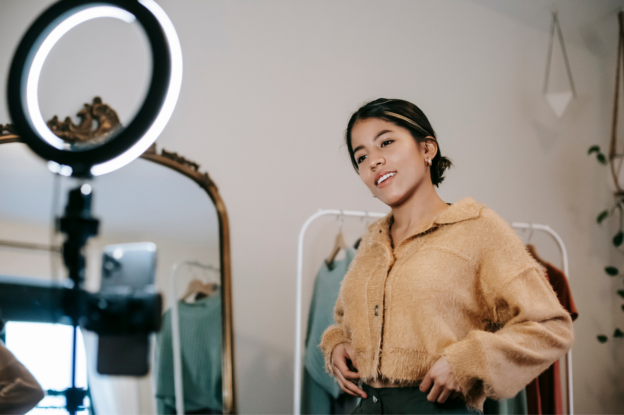 Foto de uma jovem influenciadora. Ela veste um casaco de cor marrom e está gravando um vídeo, com luzes de ringlight ligadas 