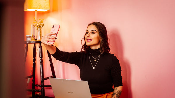 Mulher tira selfie com o celular em uma sala de estar, de frente para o laptop.